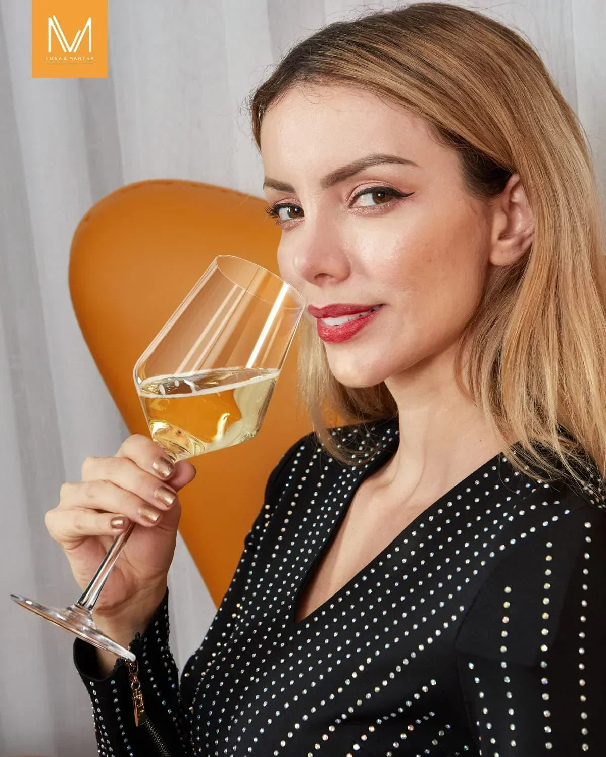 Woman sipping white wine from elegant glass, wearing black studded dress, orange chair background