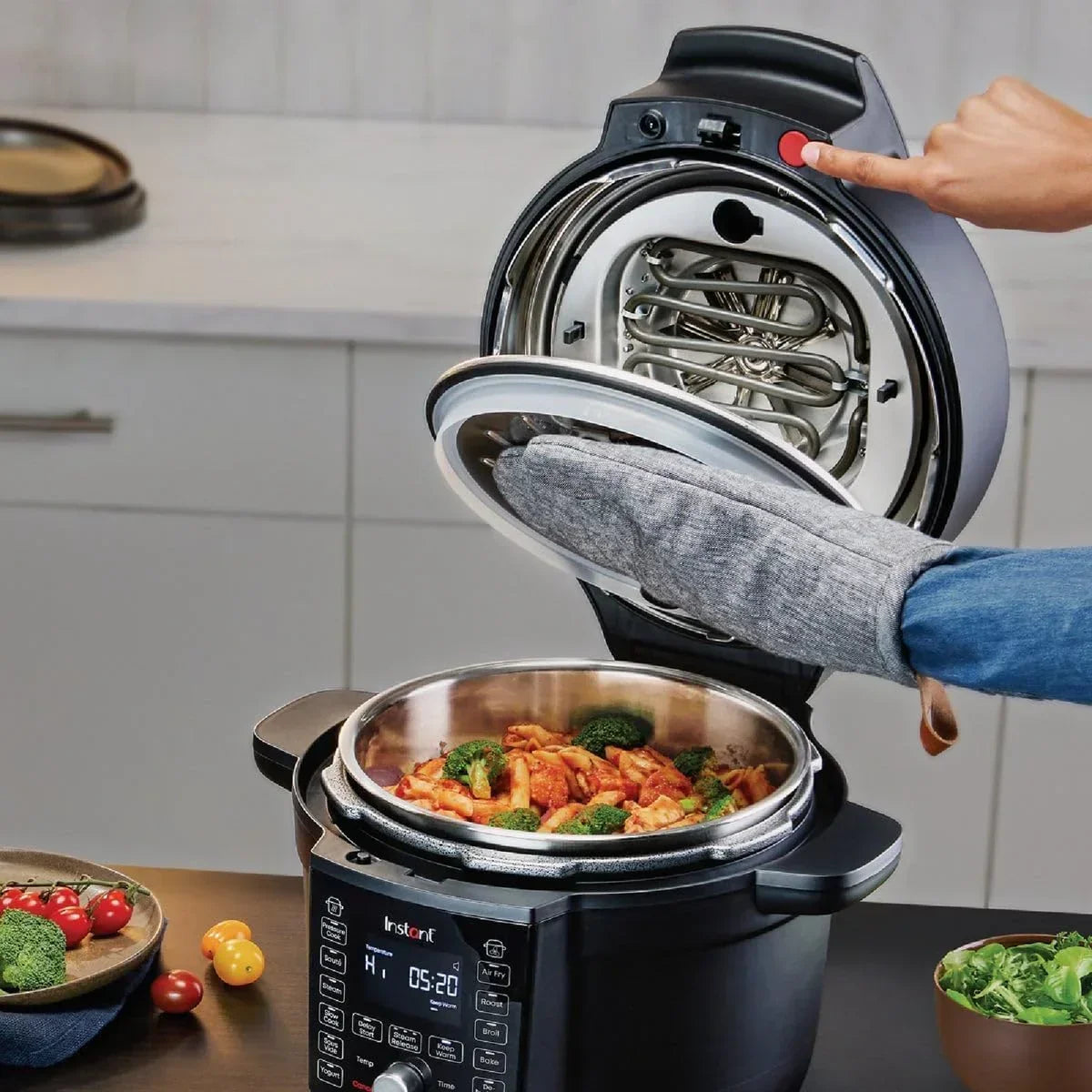 Instant Pot air fryer lid open, revealing cooked pasta with broccoli, hand pressing button, kitchen countertop