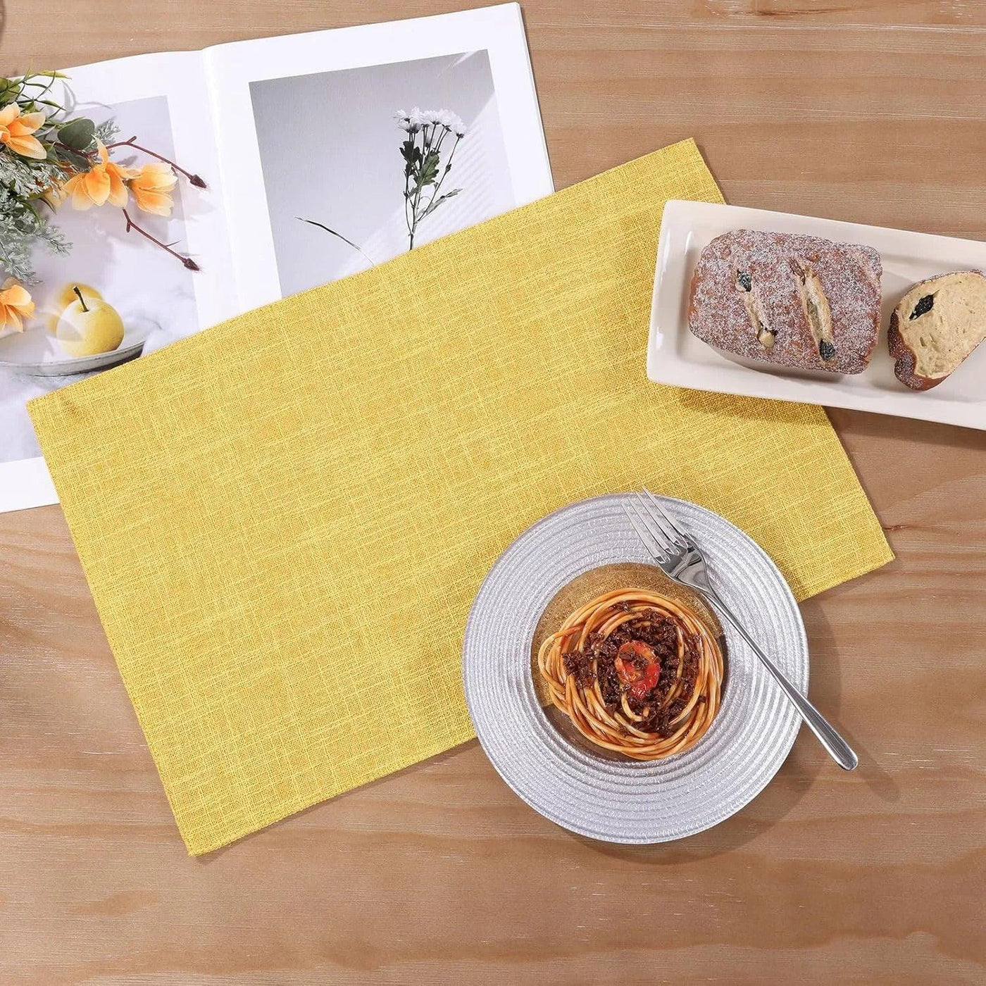 Yellow woven placemat with spaghetti plate, bread, and cookbook on wood table for kitchen dining setup