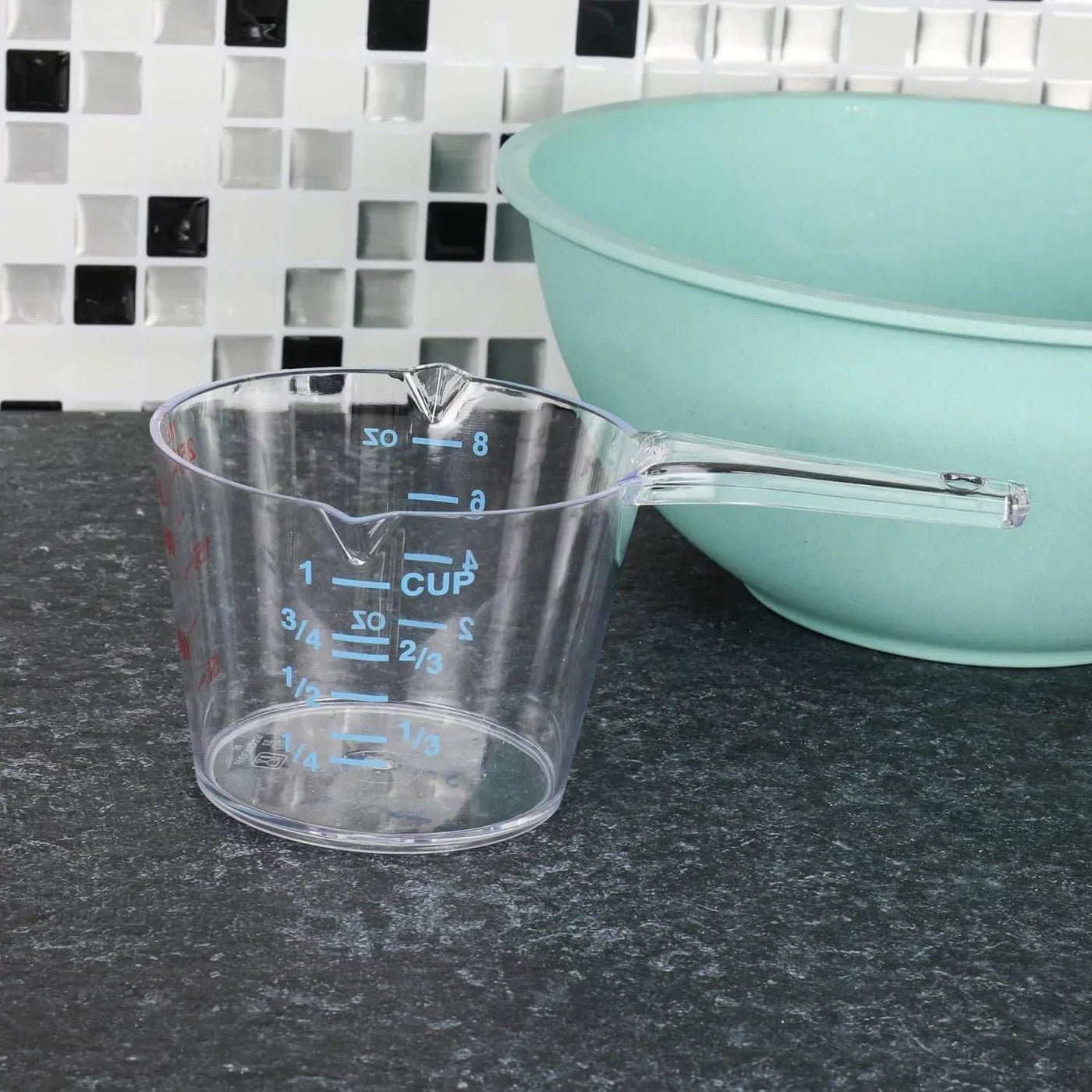 Clear plastic measuring cup with blue and red markings next to mint green mixing bowl on kitchen counter