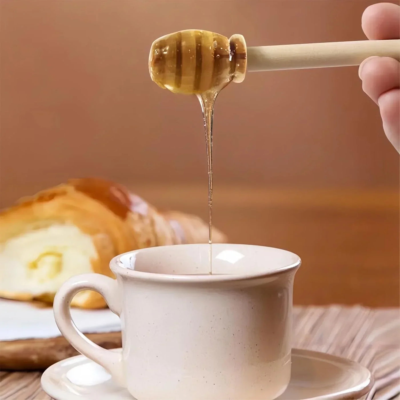 Wooden honey dipper drizzling honey into ceramic mug with croissant, kitchen breakfast scene