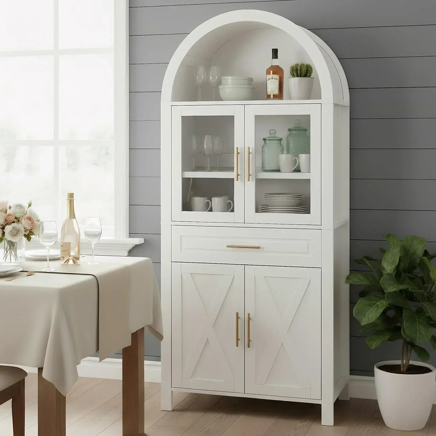 White arched kitchen storage cabinet with glass doors, gold handles, and dining table nearby