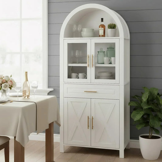 White arched kitchen storage cabinet with glass doors, gold handles, and dining table nearby