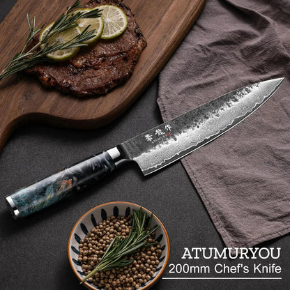 Damascus steel chef's knife with patterned blade beside steak, pepper, and rosemary on dark kitchen surface.