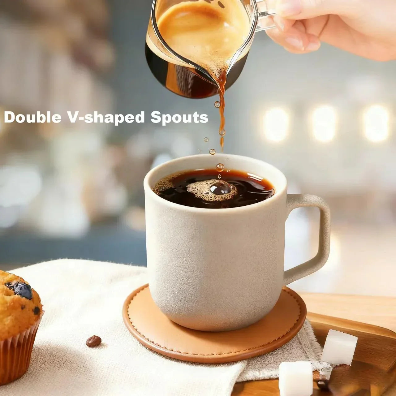 Pouring espresso from glass pitcher with double V-shaped spouts into ceramic mug, coffee setup with muffin and sugar cubes