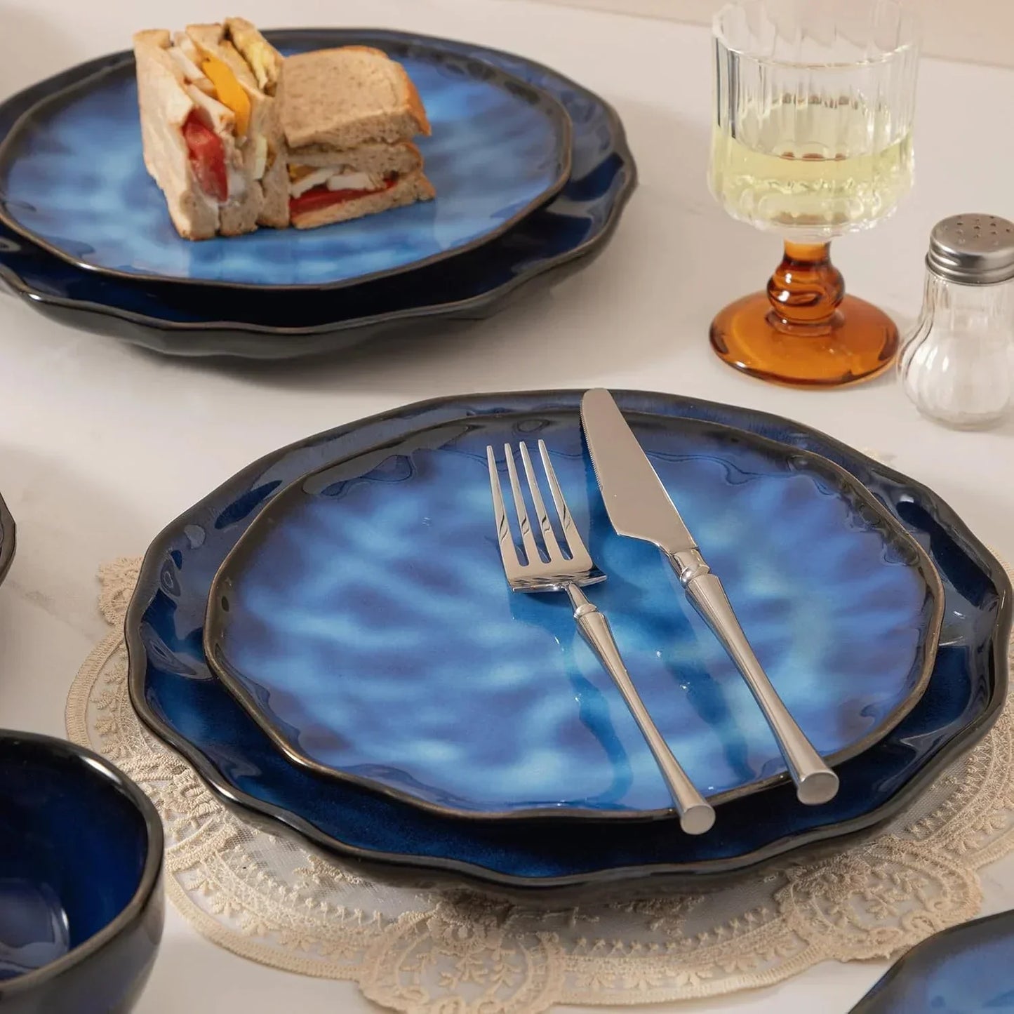 Blue ceramic dinner plates with scalloped edges, silverware, and a sandwich, set on lace placemats