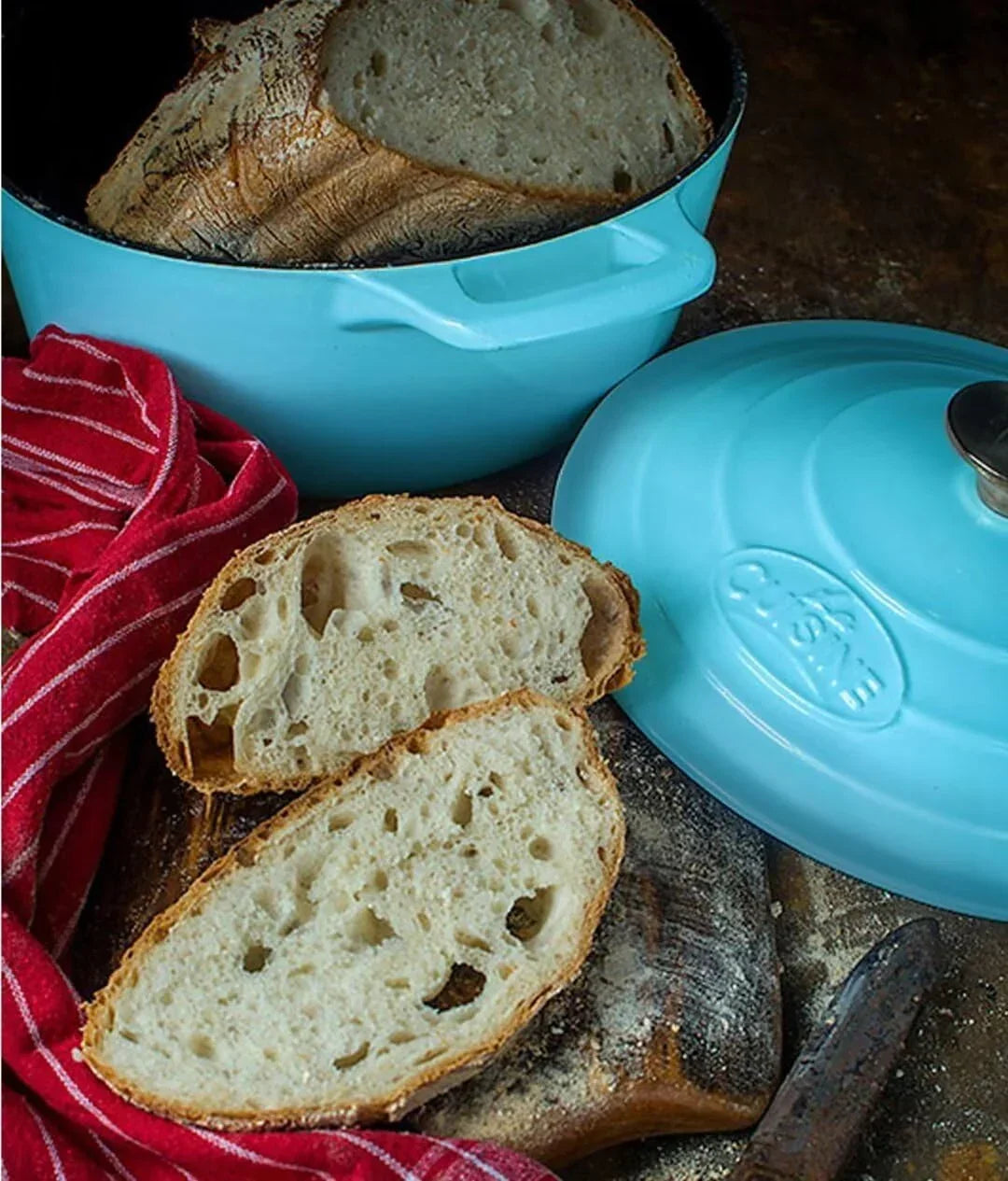 Freshly baked artisan bread in a blue Dutch oven with sliced loaf and red kitchen towel