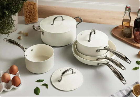 White nonstick cookware set with lids on kitchen table, surrounded by eggs, pasta, and spices