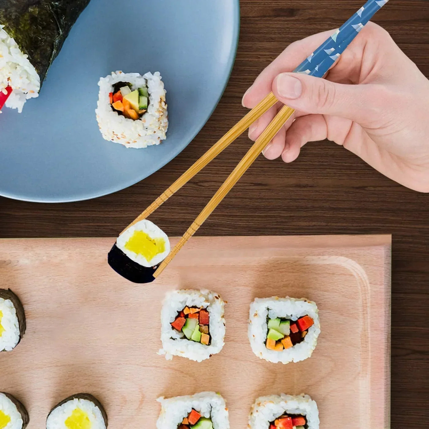 Bamboo chopsticks picking sushi roll from wooden board, blue plate with sushi nearby