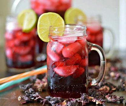 Glass mason jar mug filled with iced hibiscus tea, lime slice garnish, and dried hibiscus flowers on table