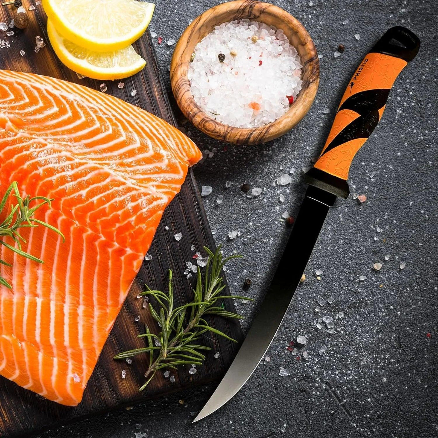 Orange-handled fillet knife beside fresh salmon, rosemary, lemon, and sea salt on dark board