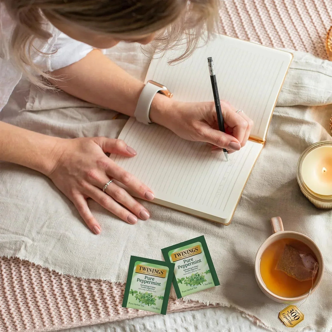 Woman writing in notebook with Twinings Pure Peppermint tea bags, cup of tea, and candle nearby