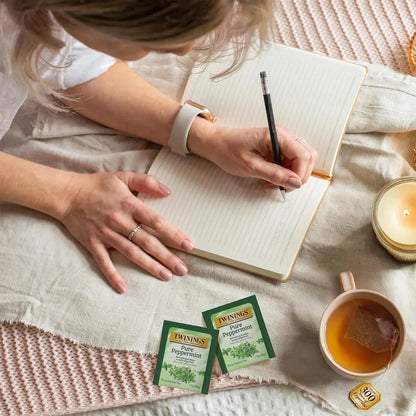 Woman writing in notebook with Twinings Pure Peppermint tea bags, cup of tea, and candle nearby