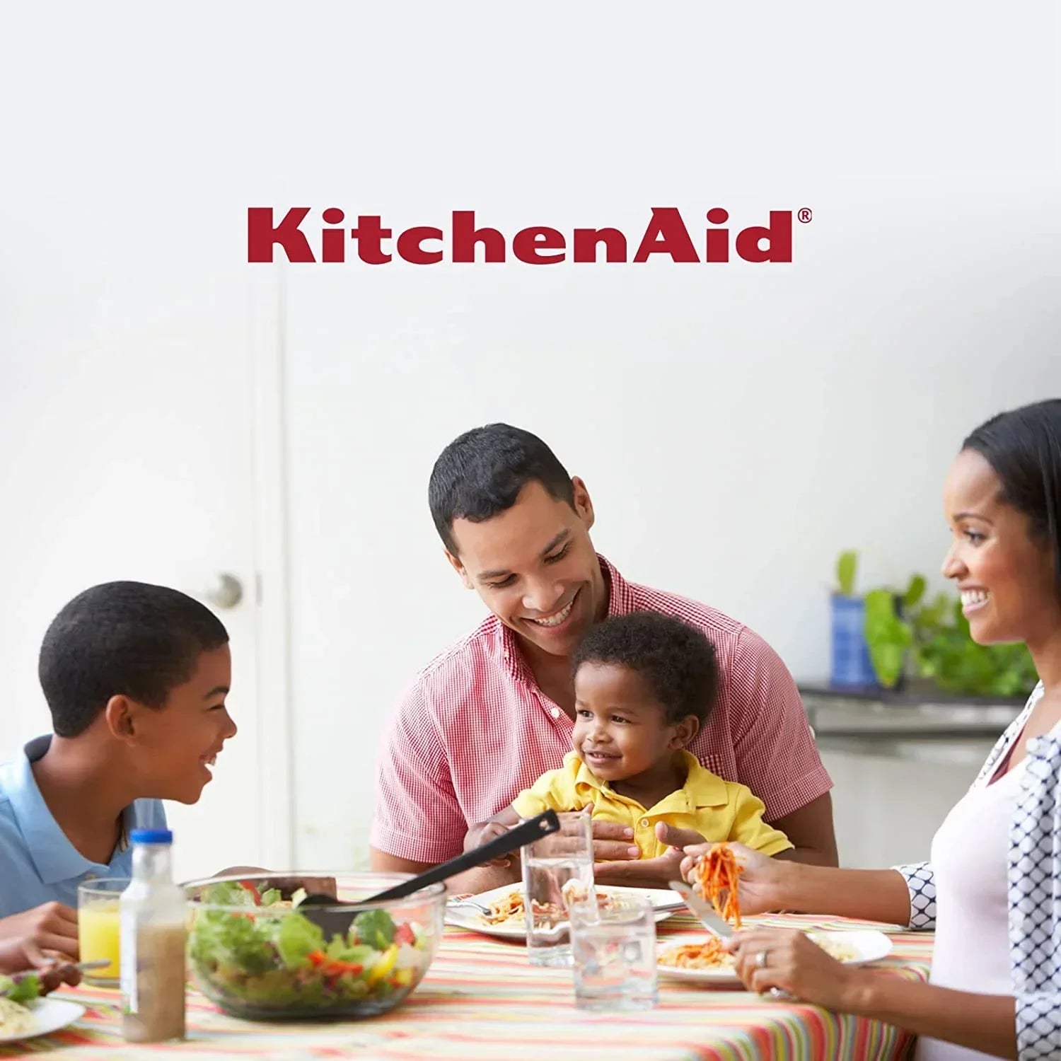 Family enjoying meal together with KitchenAid logo, featuring pasta, salad, and tableware in bright kitchen setting
