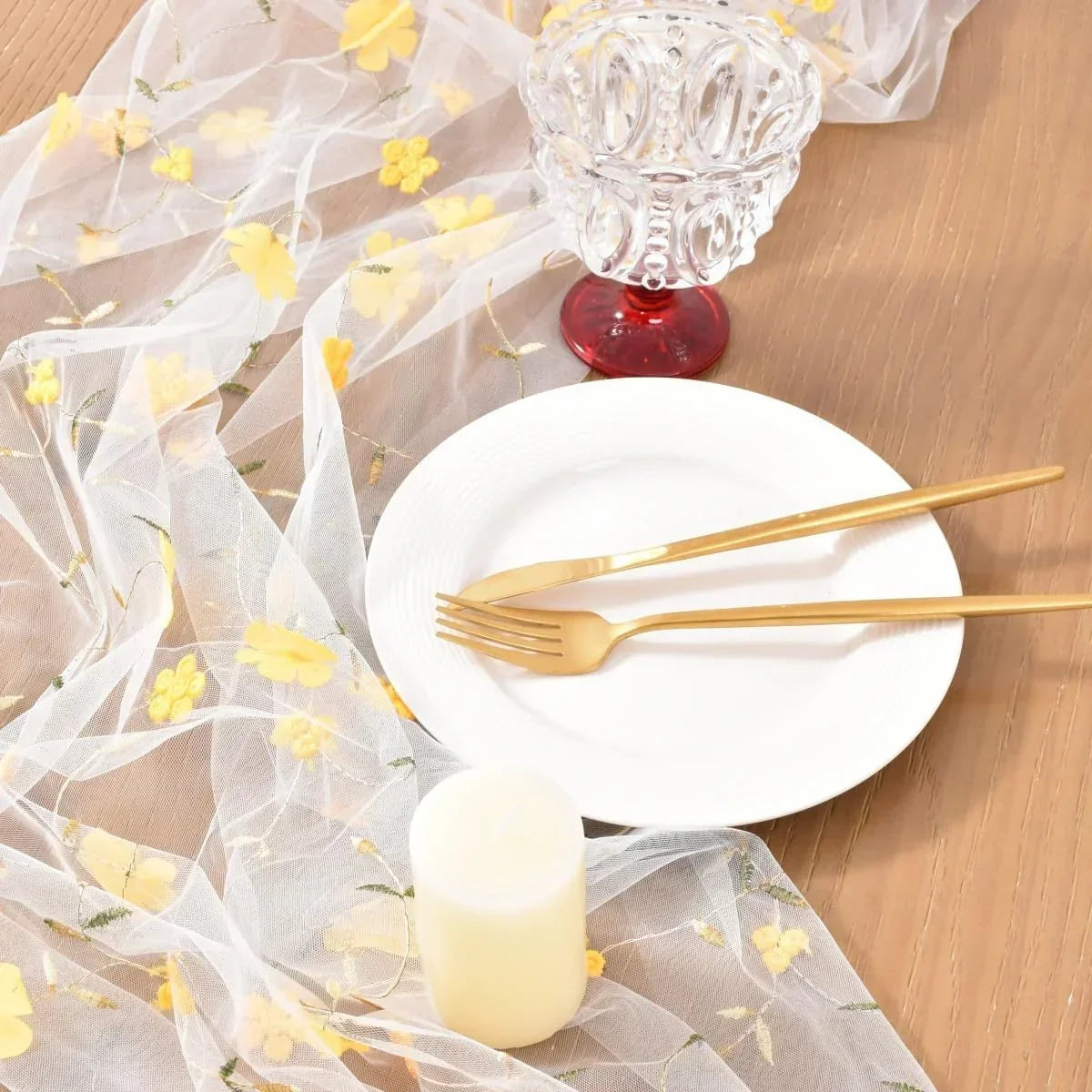 White plate with gold fork and knife, floral sheer table runner, crystal glass, and candle on wood table