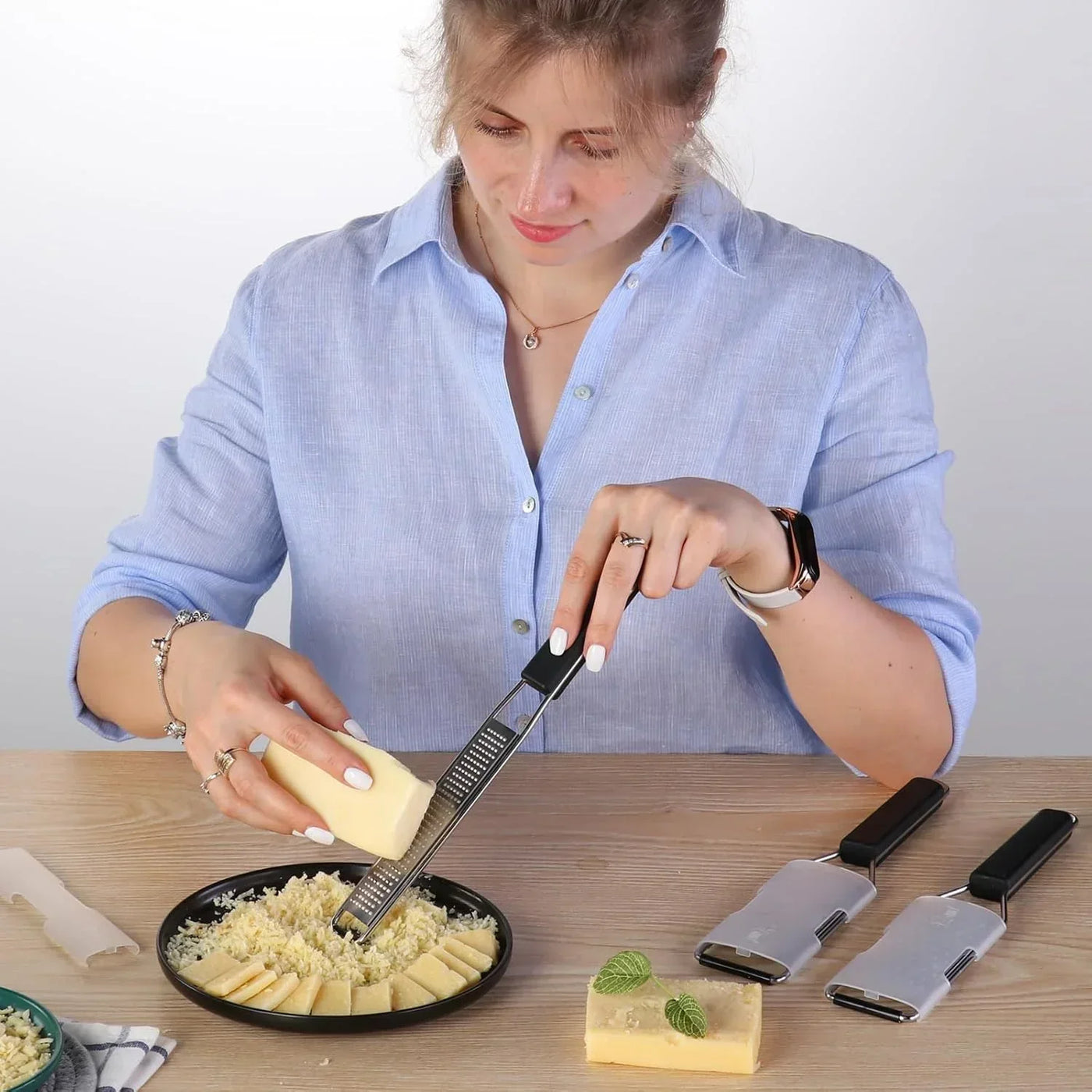 Woman grating cheese with kitchen grater, cheese slices on plate, kitchen tools on wooden table
