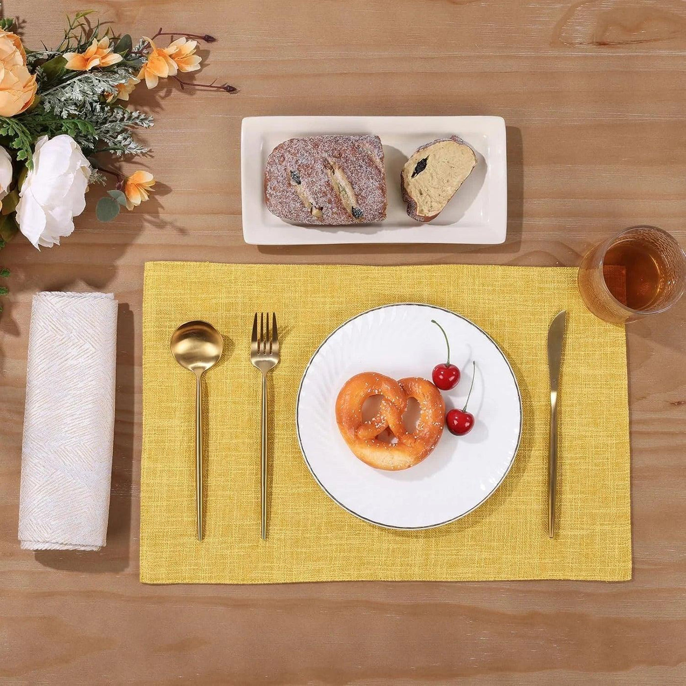 Yellow woven placemat with gold cutlery, pretzel, cherries, and bread on wood table for dining setup