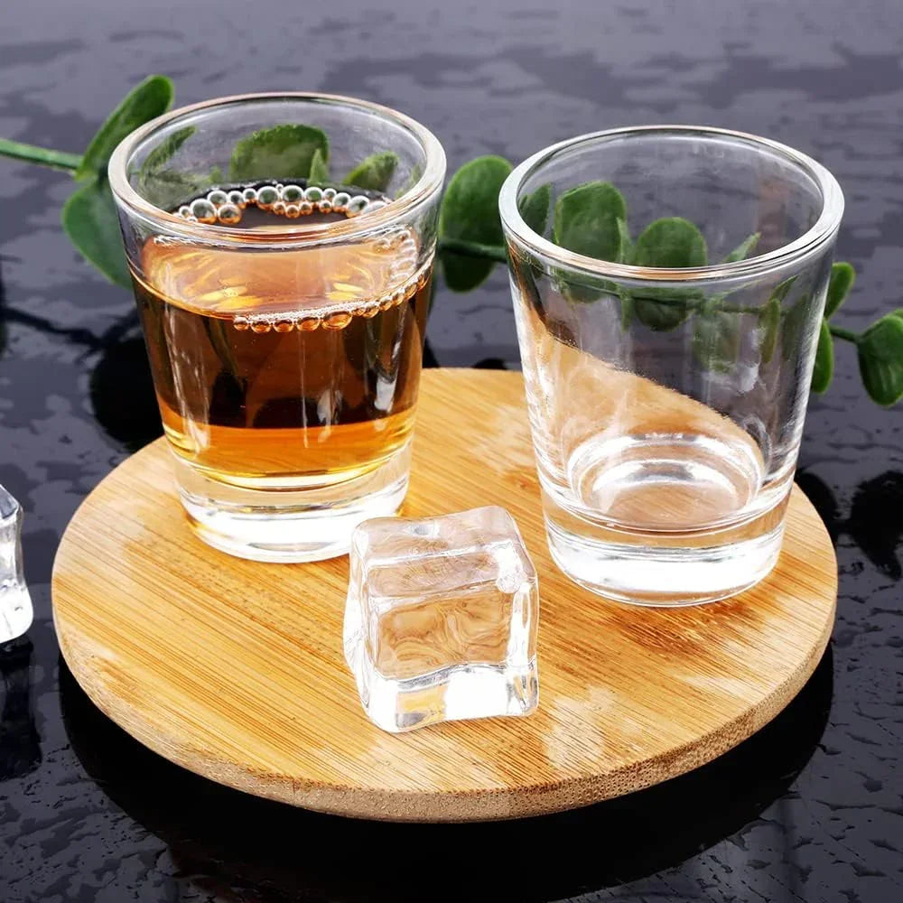 Clear glass shot glasses with amber liquid and ice on bamboo coaster, green leaves background