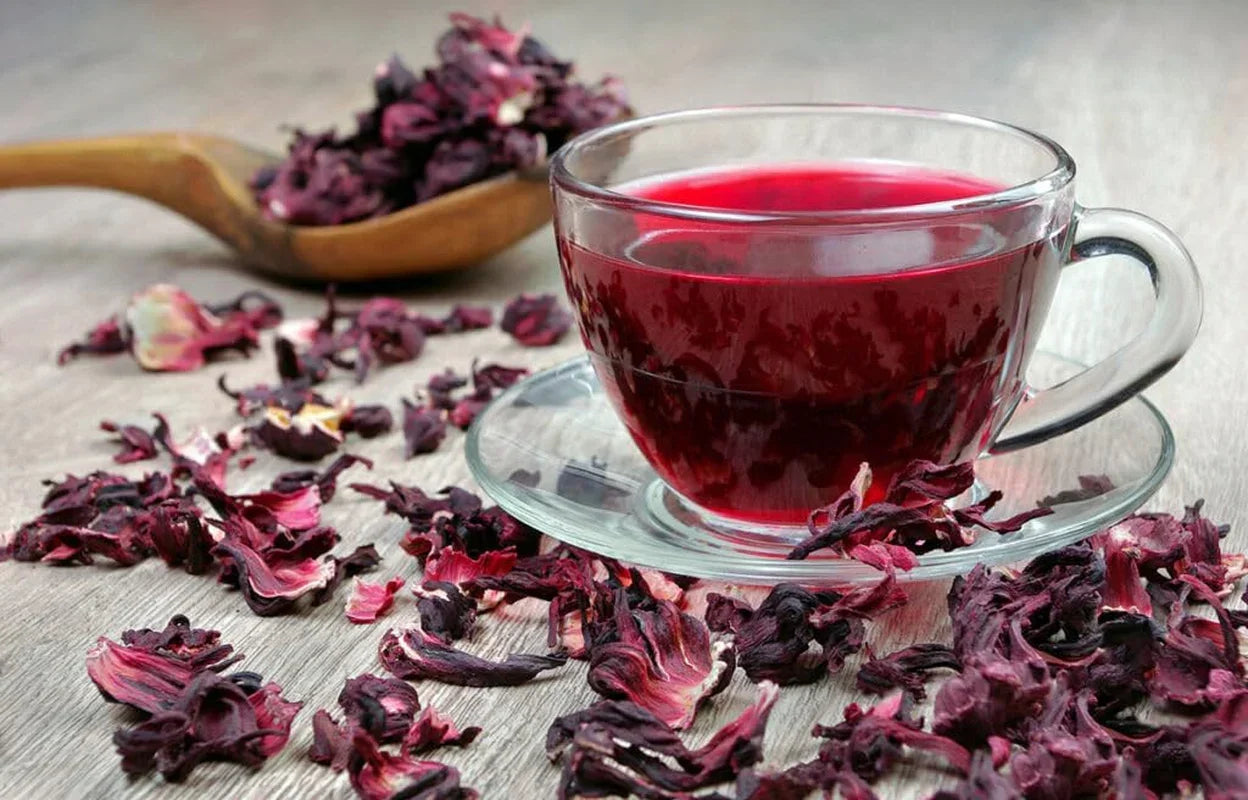 Clear glass teacup with vibrant red hibiscus tea, surrounded by dried hibiscus flowers on wooden surface
