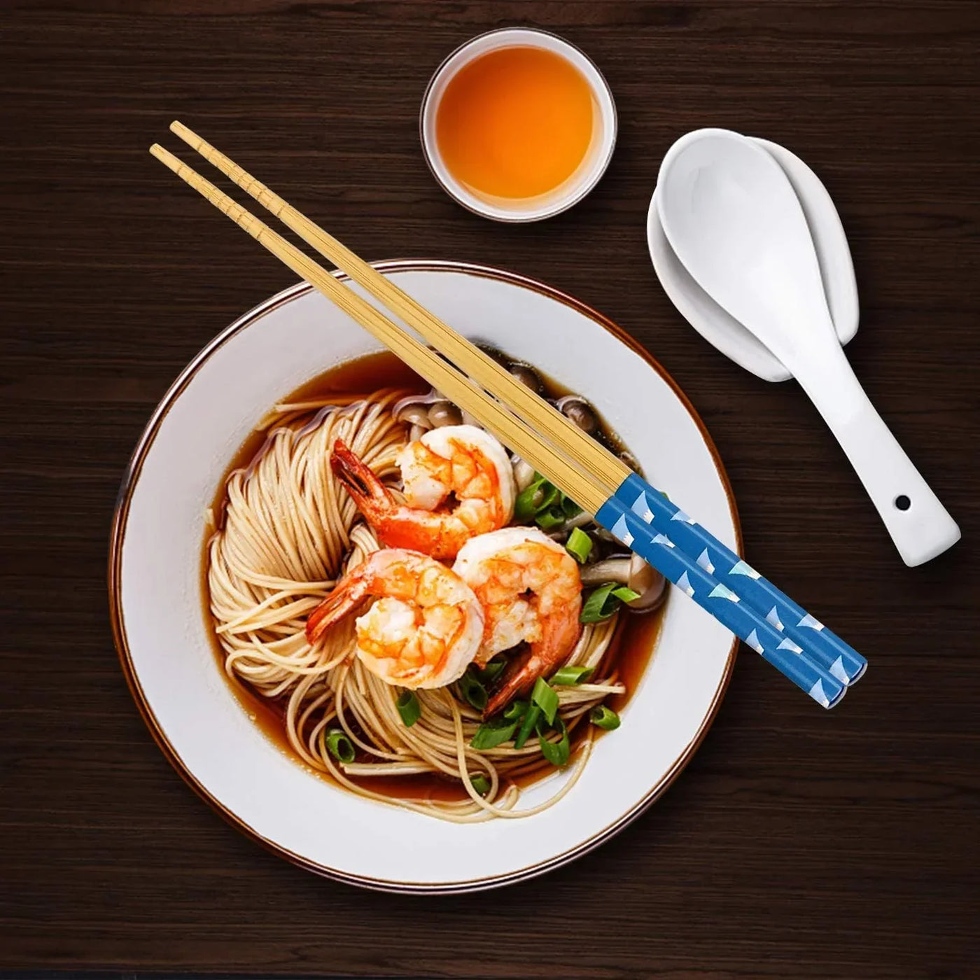 Bamboo chopsticks with blue pattern, shrimp noodle bowl, white spoons, and tea on dark wood table