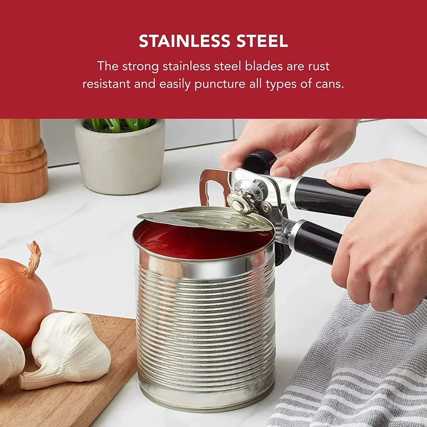 Stainless steel can opener opening tomato can with garlic, onion, and kitchen towel nearby